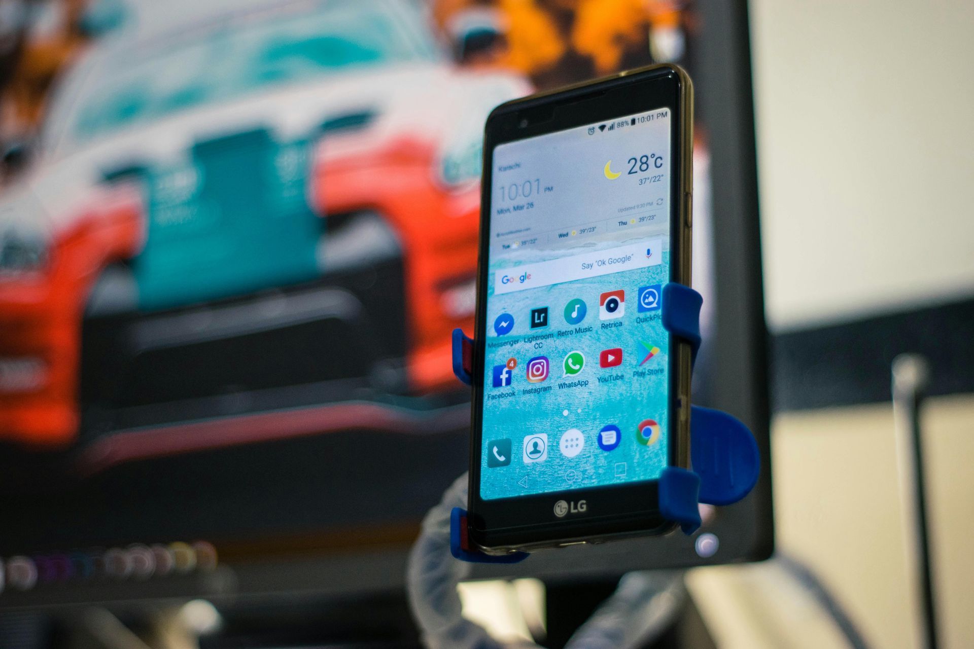 LG-Smartphone in einer blauen Halterung, auf dem Apps angezeigt werden, vor einem farbenfrohen Rennwagen-Hintergrund.