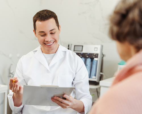 Doctor in white coat smiles while holding clipboard, speaking to a patient in a medical office.