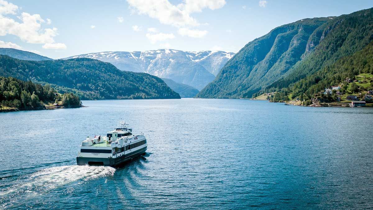 Passenger ferry sailing on blue fjord waters between green, mountainous shores. Snowy peaks in the distance, blue sky.