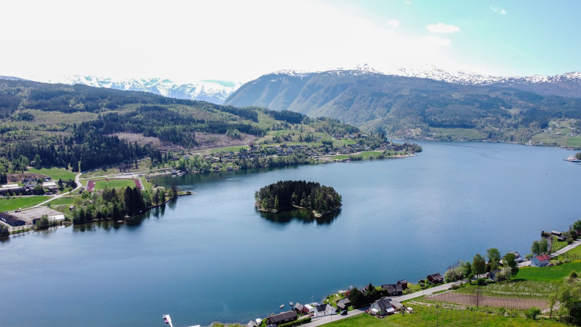 A scenic landscape with a fjord, island, surrounding forest, and snow-capped mountains in Ulvik, Hardangerfjord.