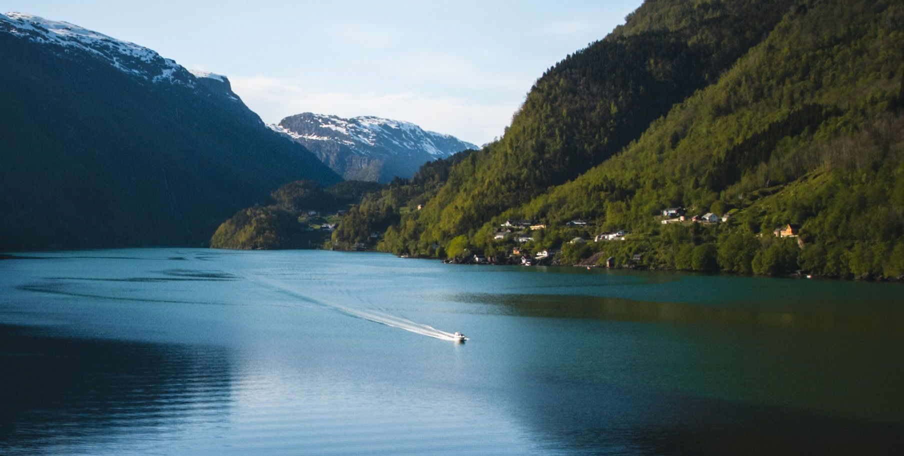 A boat travels across a fjord, flanked by lush green mountains under a blue sky.