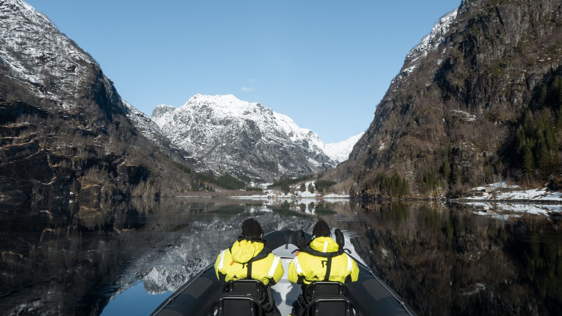 Two people in yellow jackets on a boat navigate a fjord, flanked by snow-capped mountains.