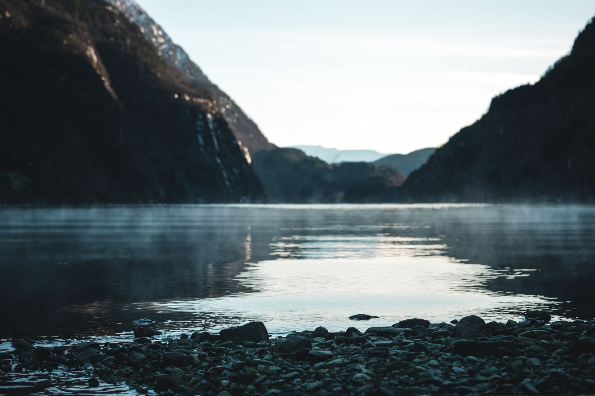 Calm fjord between dark mountains; fog hovers above water; sunny sky.