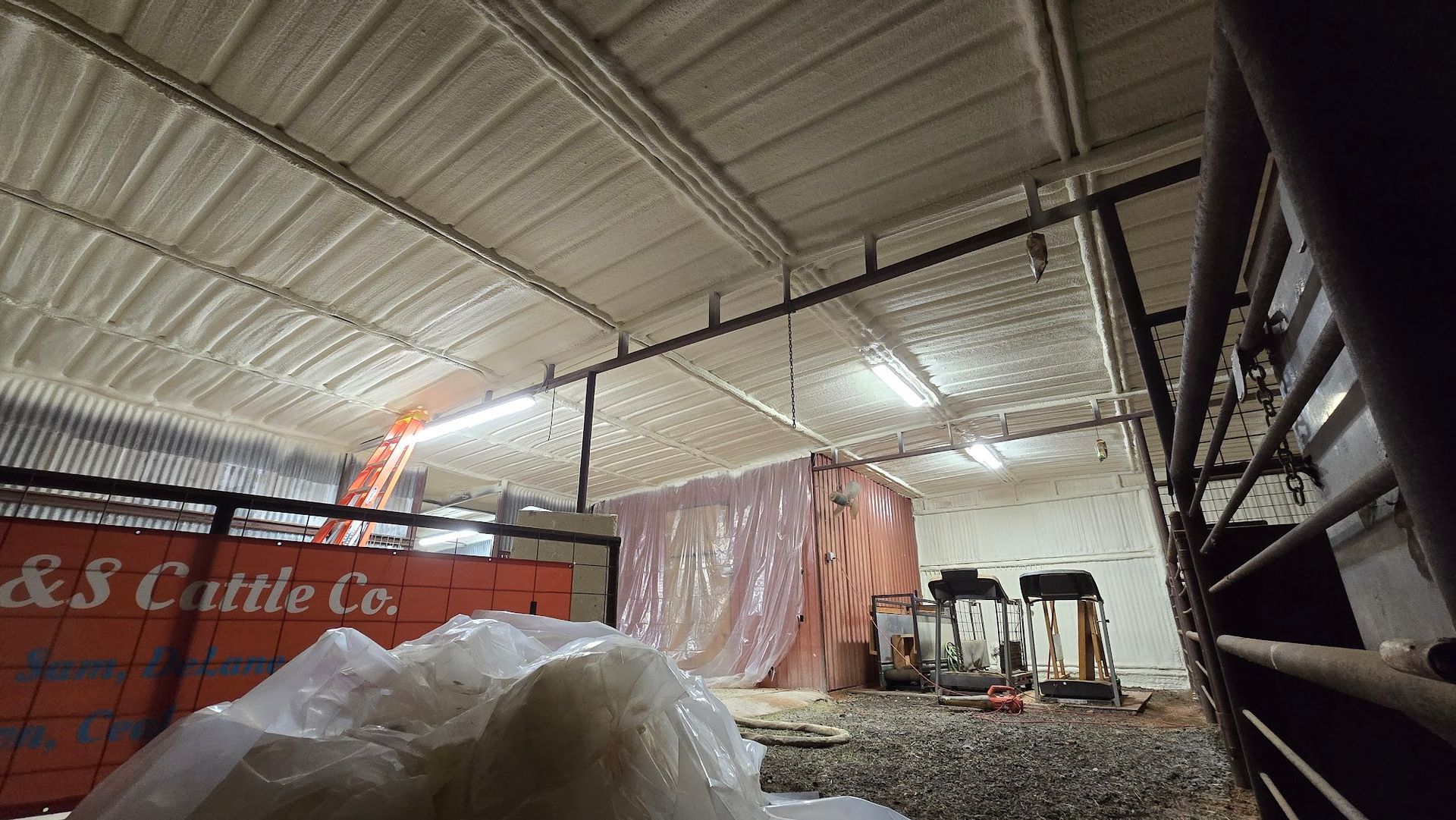 Inside a barn, a covered vehicle and equipment sit under a metal roof beside a tarp-covered pile.
