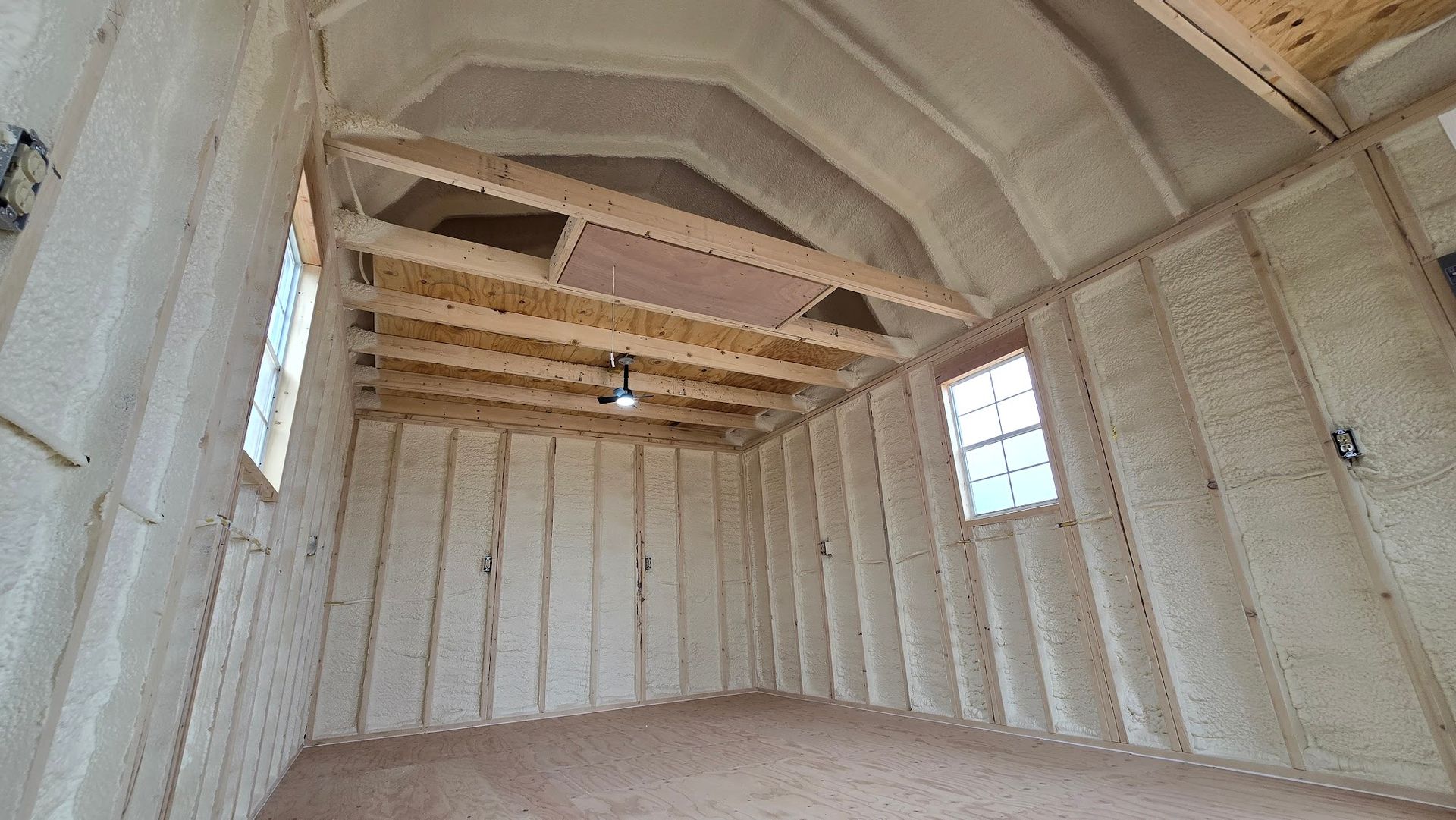 Unfinished insulated room with exposed ceiling beams and two small windows.