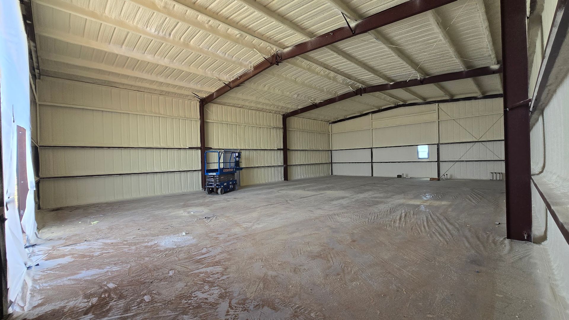 Empty metal warehouse interior with exposed beams, dirt floor, and a blue forklift near the back wall