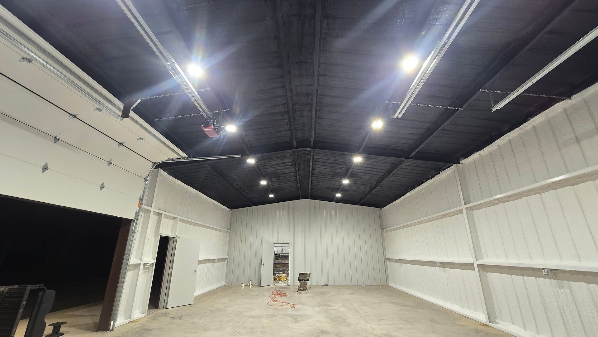 Empty warehouse interior with high black ceiling, bright lights, and unfinished white walls and concrete floor