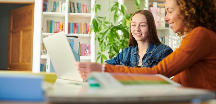 A mom and daughter are both sitting at a table, smiling and look at a laptop screen