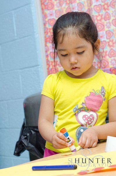 A Little Girl in A Yellow Shirt Is Sitting at A Table Holding a Glue Stick.