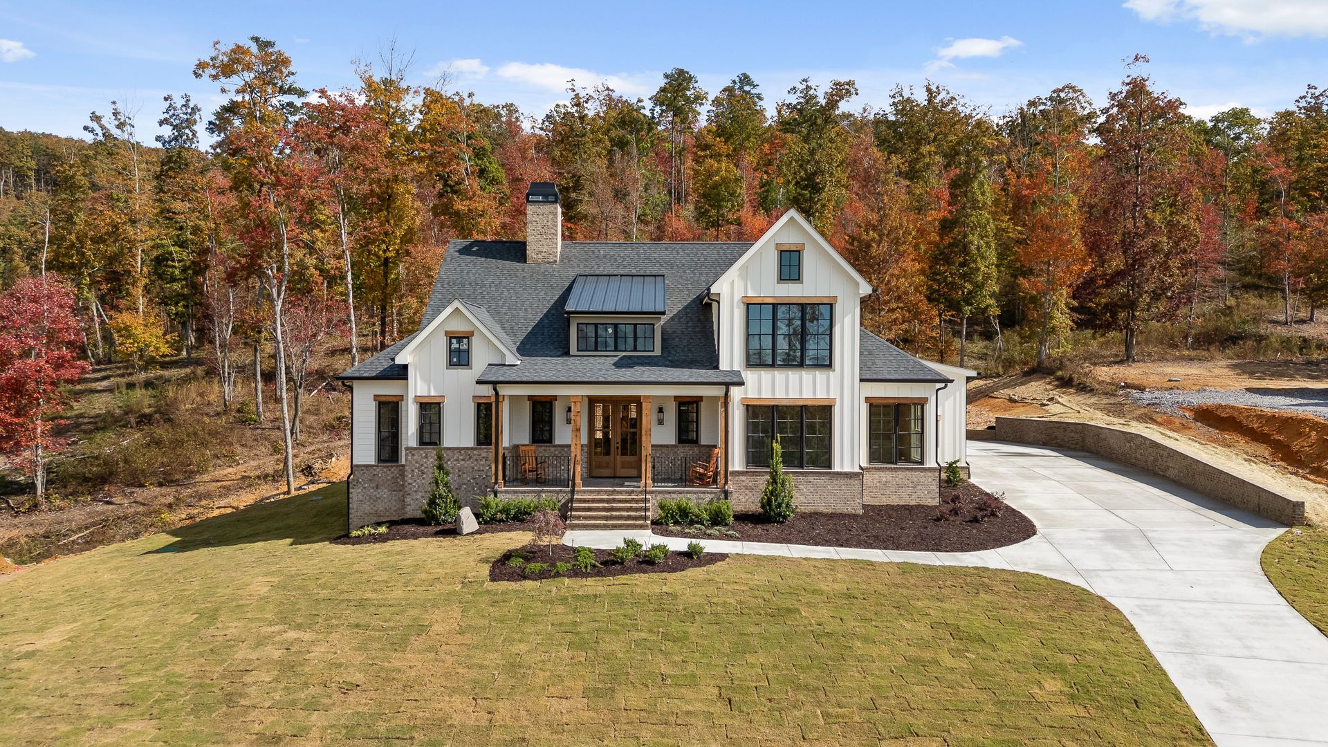 Large white farmhouse with gray roof and wraparound porch, set on a lawn beside an autumn tree line.