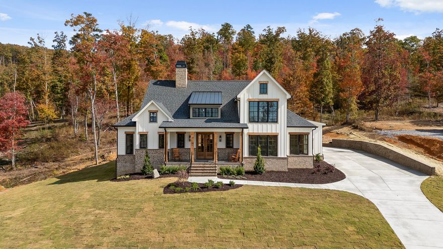 A custom home in River Gorge Ranch from NuHaven Homes that features a Large white farmhouse with gray roof and wraparound porch, set on a lawn beside an autumn tree line.
