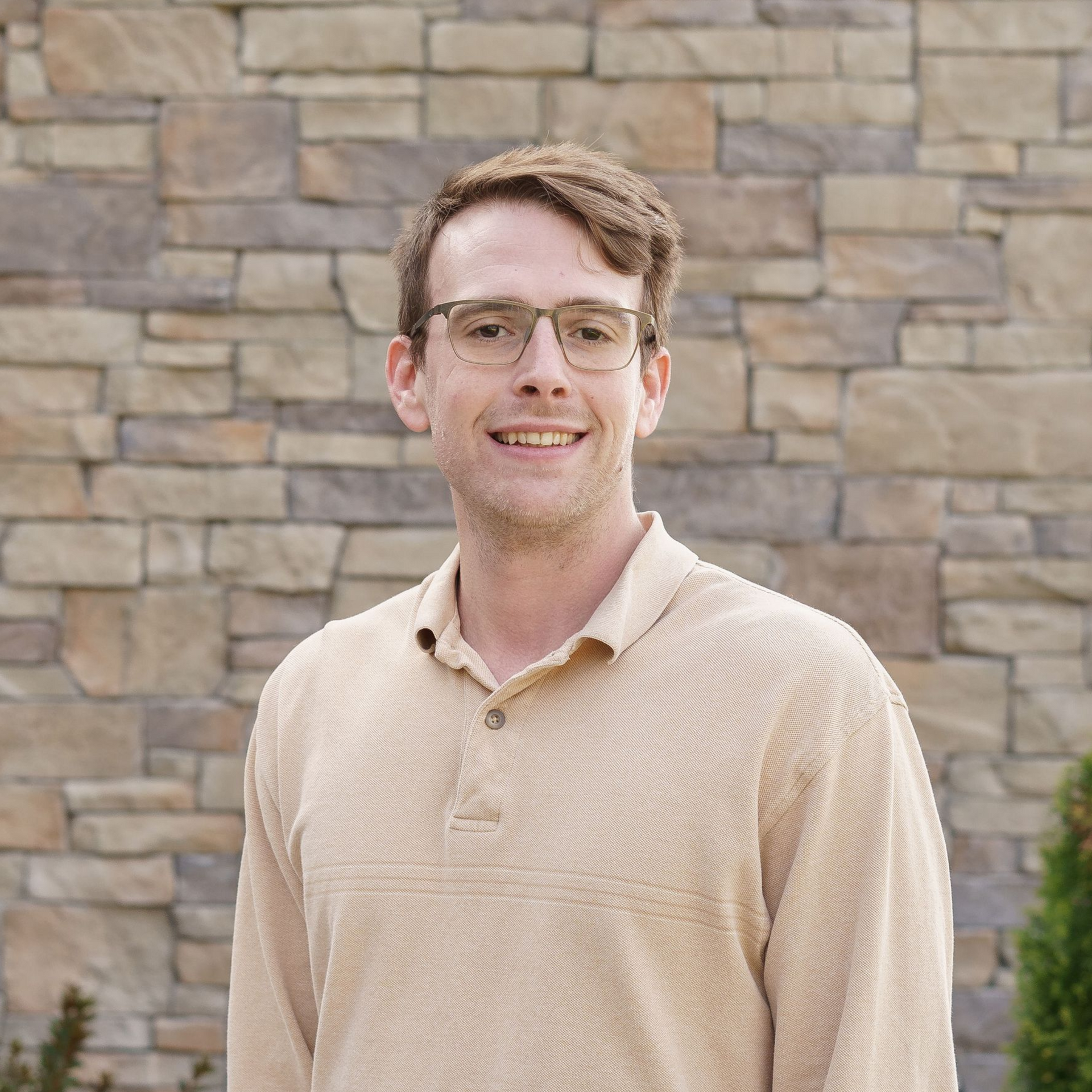 Man smiling in a maroon shirt and black vest, standing before a stone wall.