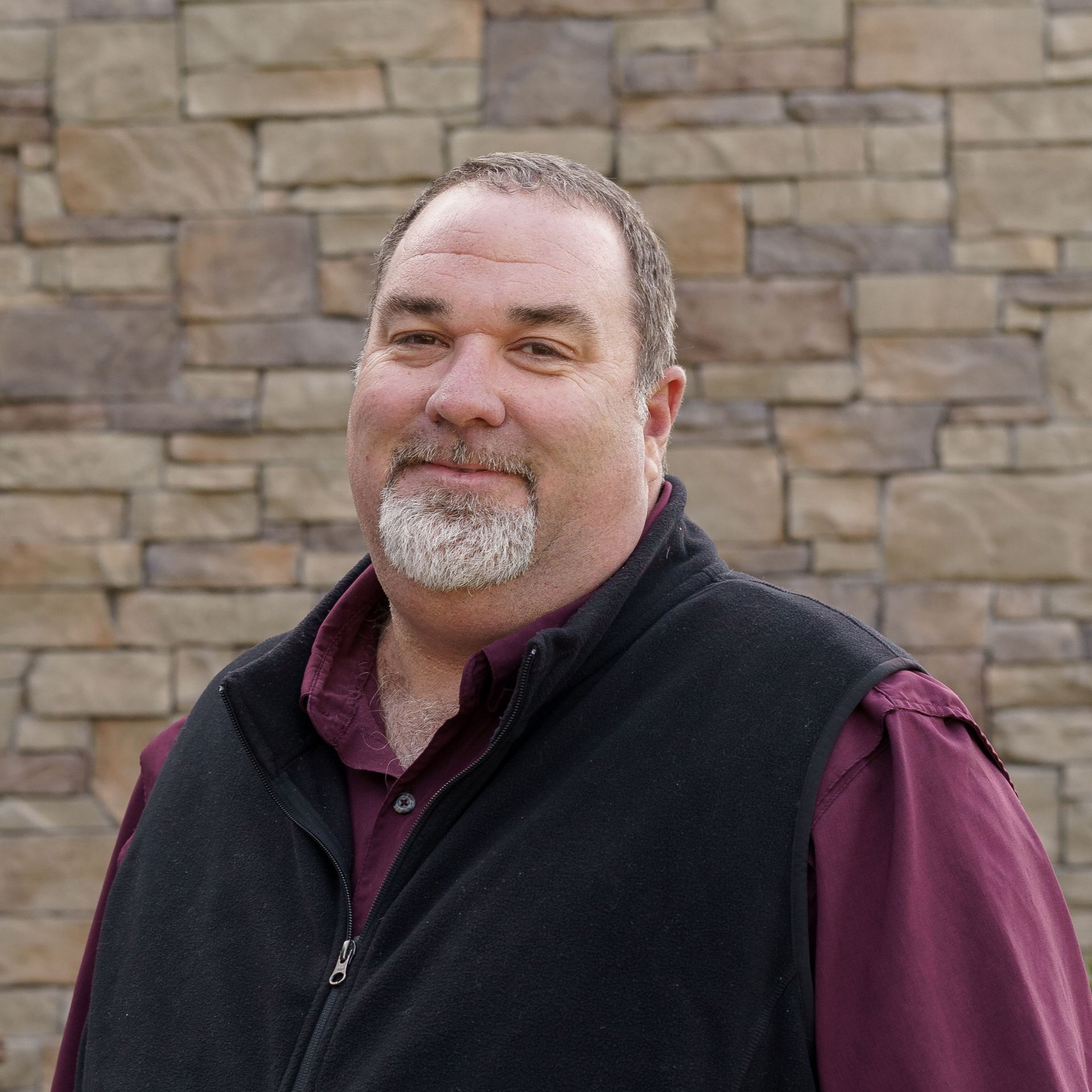 Man with red hair and beard standing before a stone wall, wearing a tan shirt and olive quilted vest.