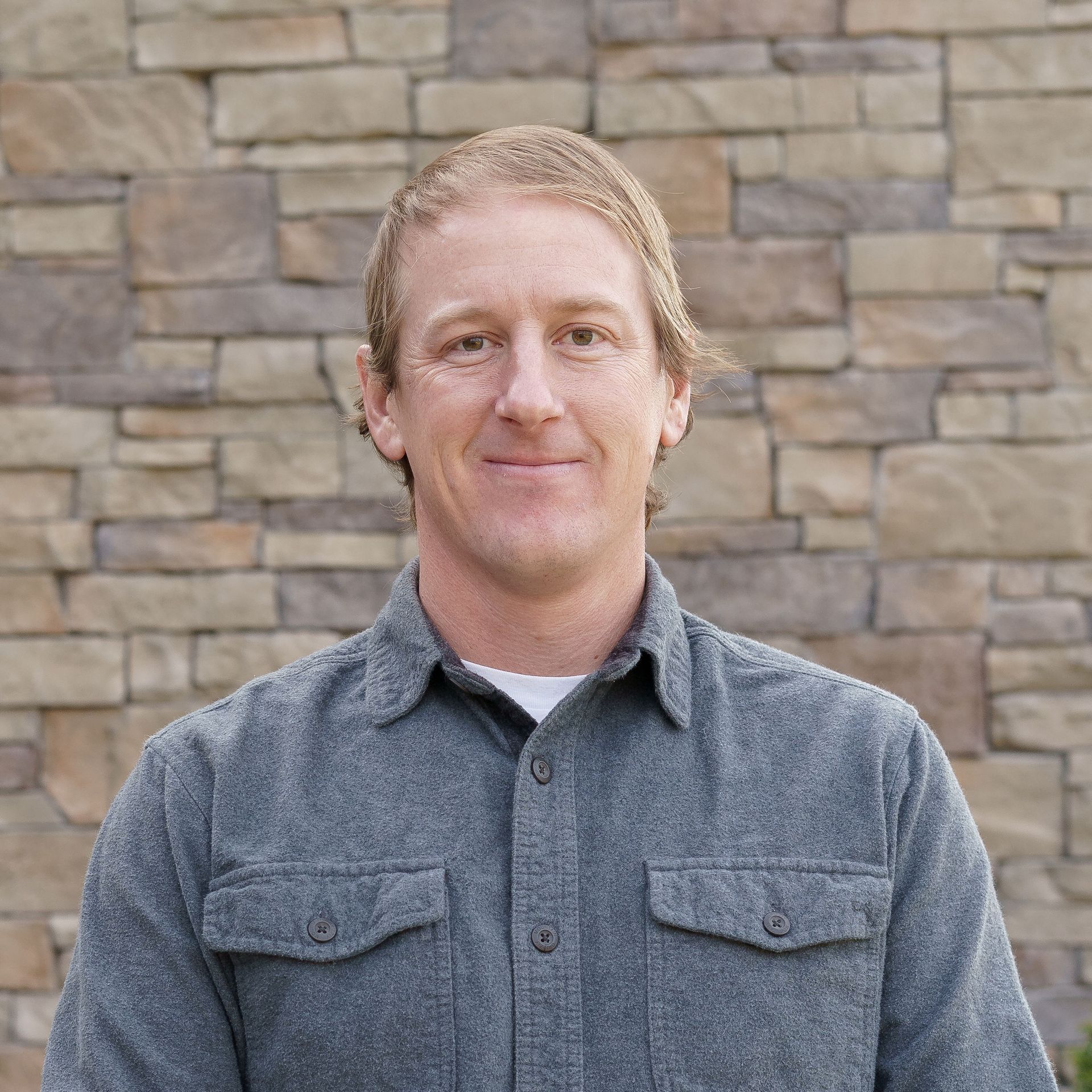 Man smiling in a maroon shirt and black vest, standing before a stone wall.