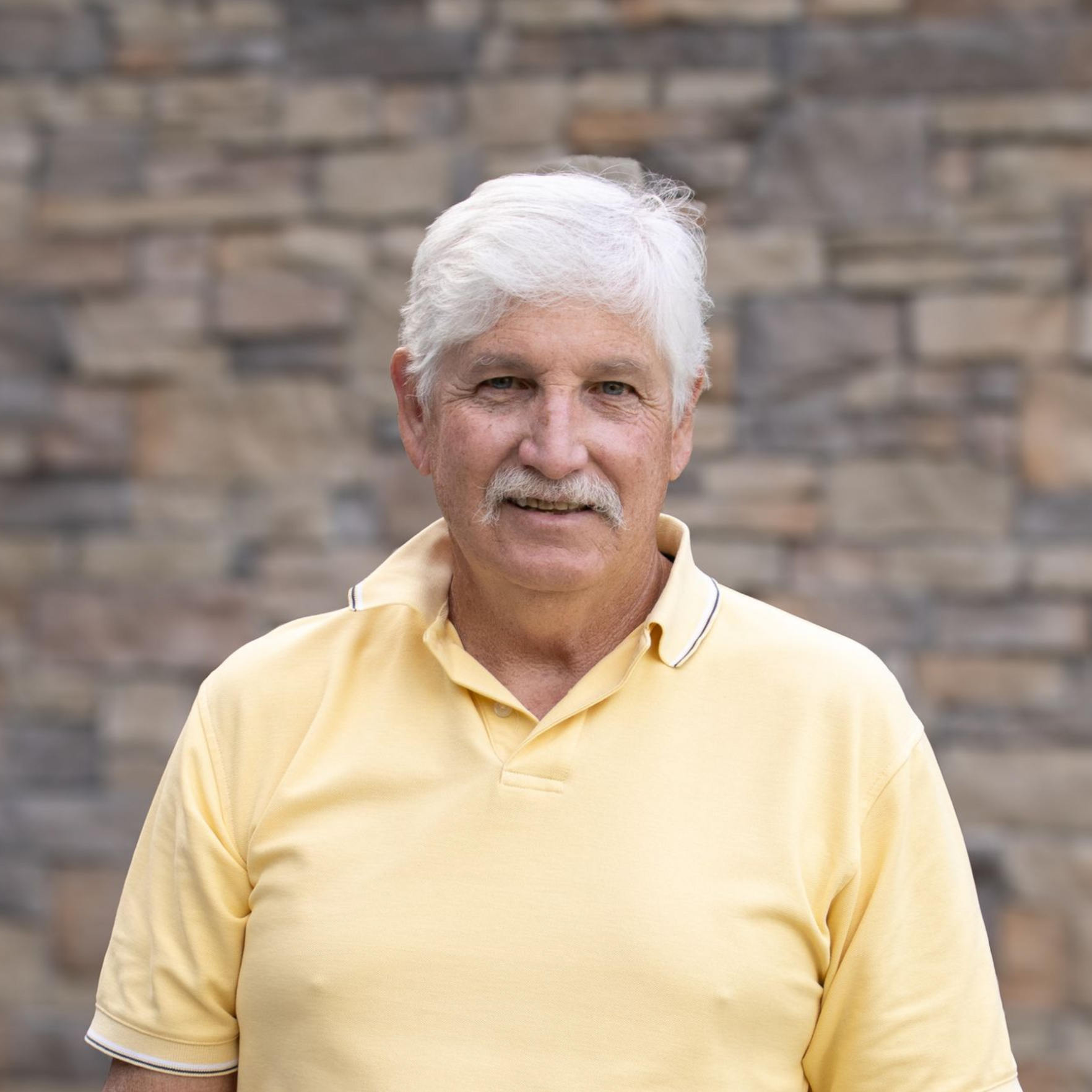 Man smiling in a maroon shirt and black vest, standing before a stone wall.
