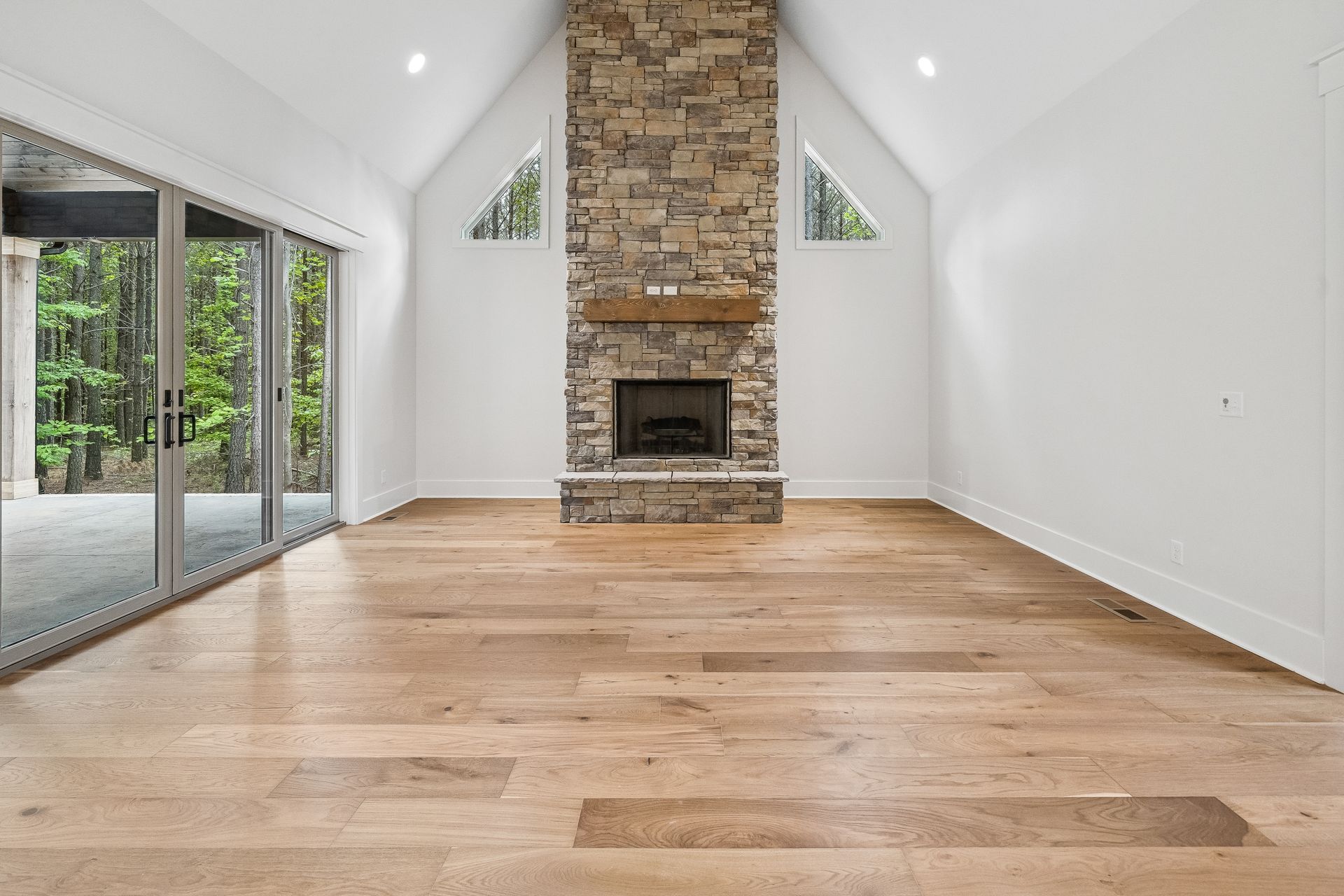 Empty living room with vaulted ceiling, stone fireplace, hardwood floors, and sliding glass doors to a patio