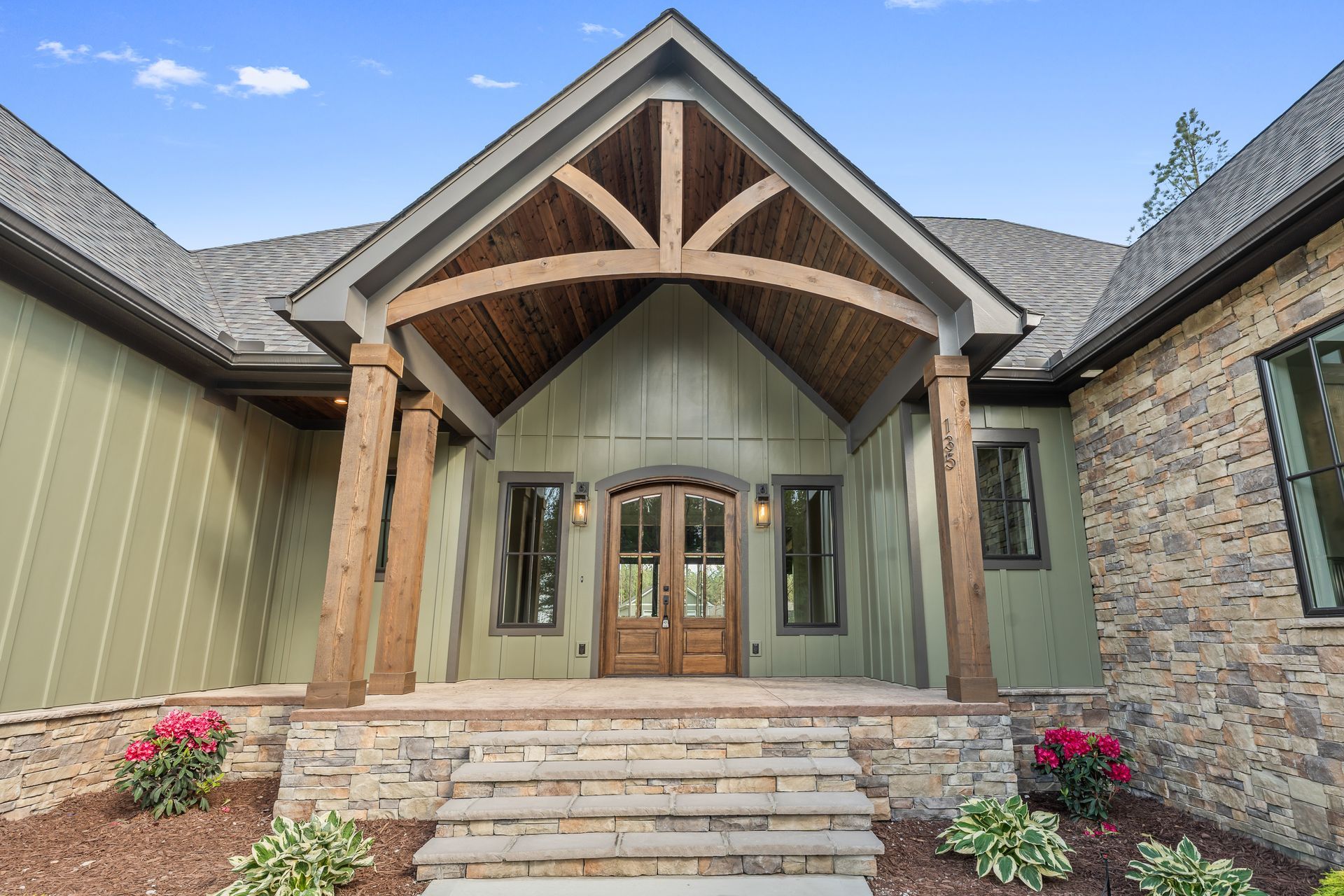 Front entrance of a modern house with timber beams, stone steps, and green siding under a peaked porch
