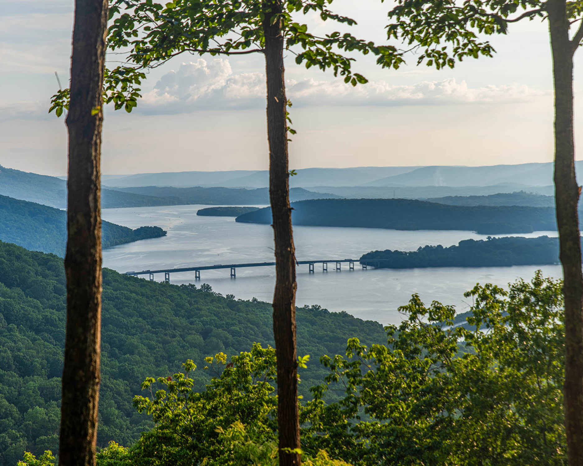 Scenic lake and bridge viewed through trees, with layered hills under a partly cloudy sky