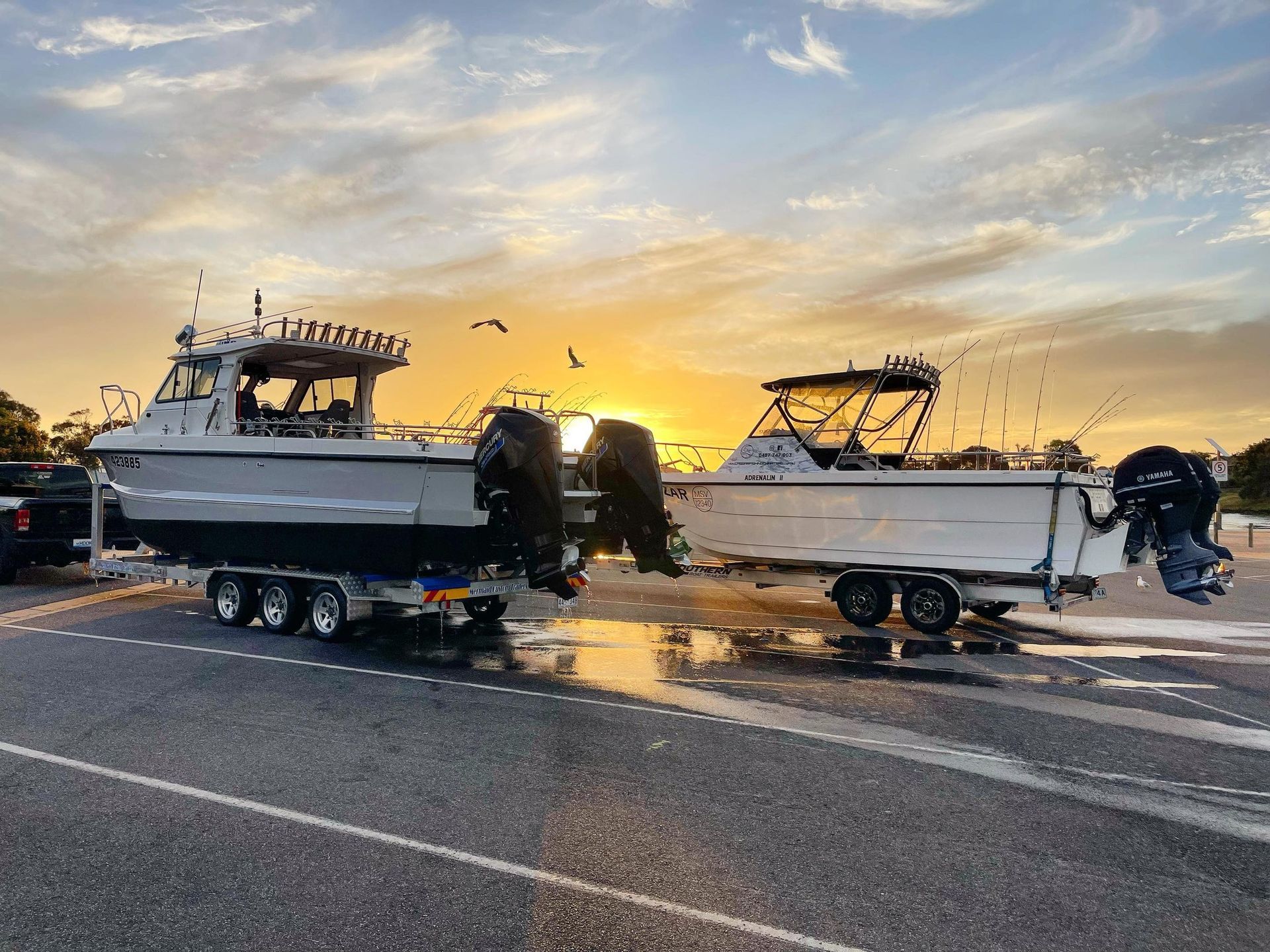 Two fishing boats on trailers at sunset in a wet parking lot with birds overhead