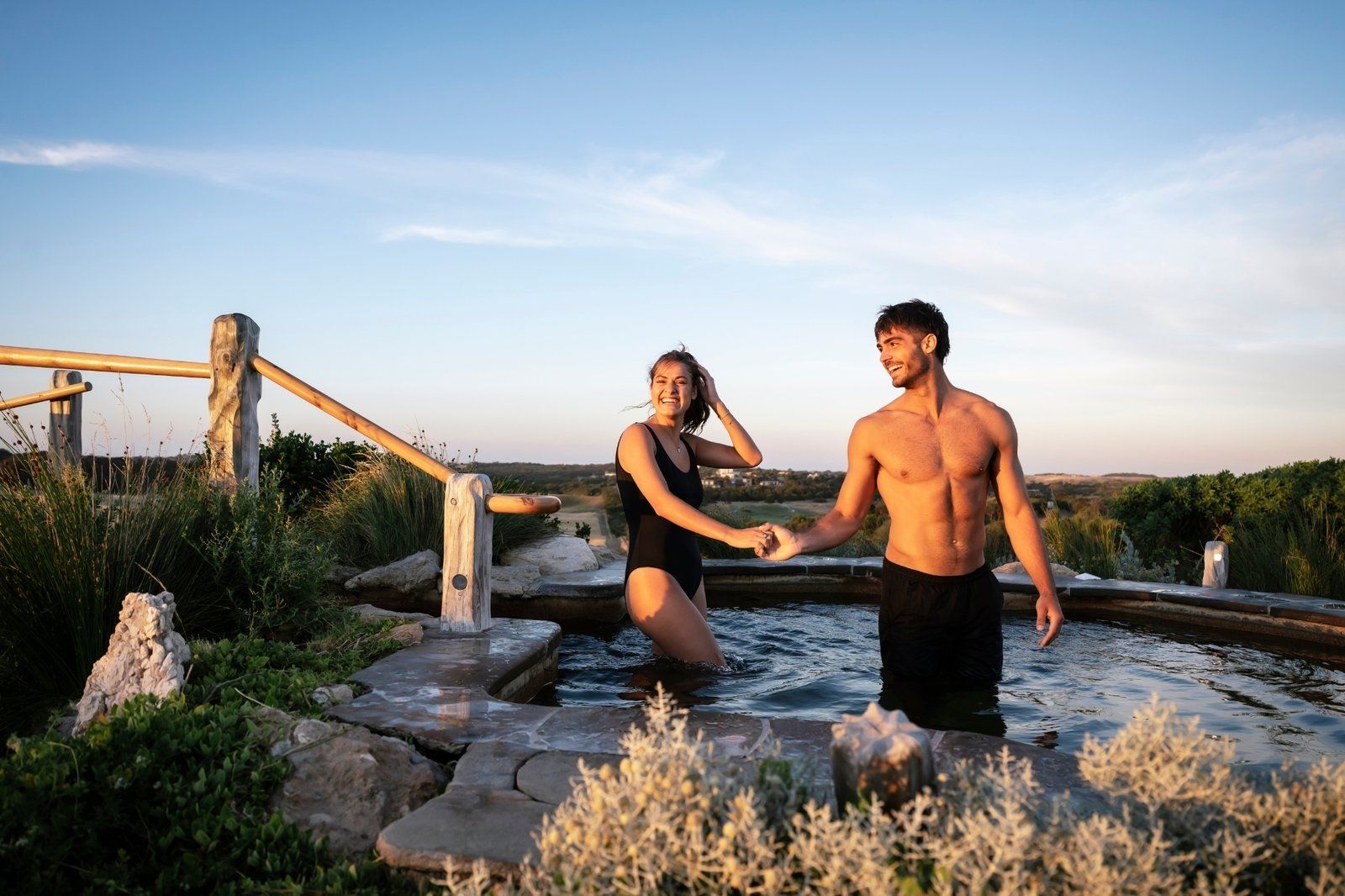 Two people standing in ocean water by a wooden walkway at sunset