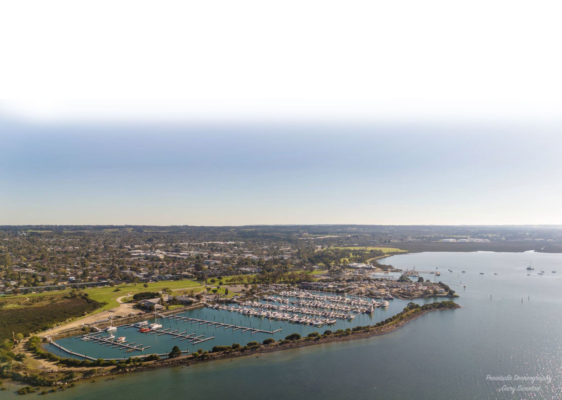 Aerial view of a coastal marina with turquoise water, boats, and a dense city skyline under a hazy sky