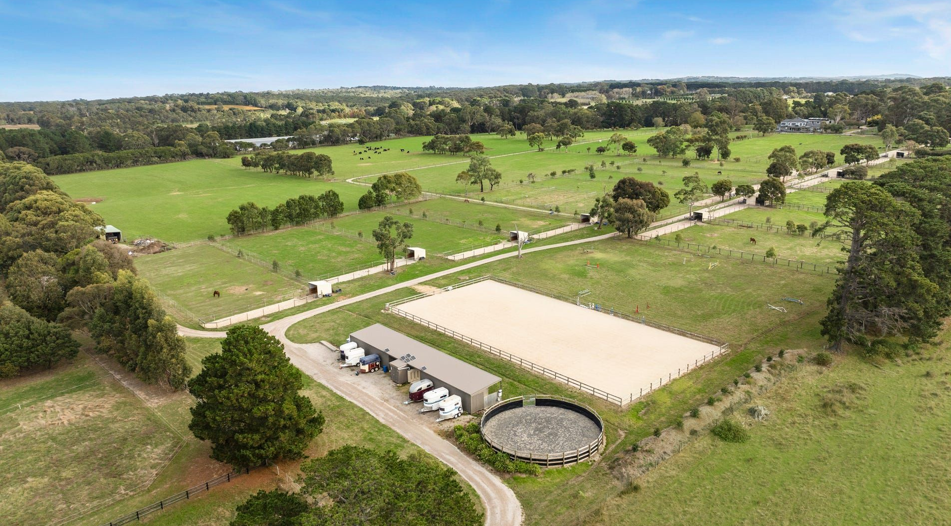 Aerial view of a grassy park with a circular pond, dirt roads, scattered trees, and a white-roofed structure