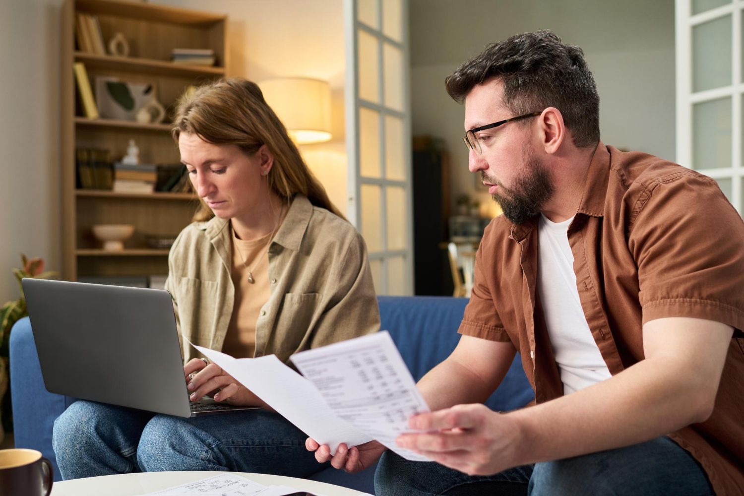 Woman using a laptop while a man looks at documents; they're sitting together on a blue couch indoors.