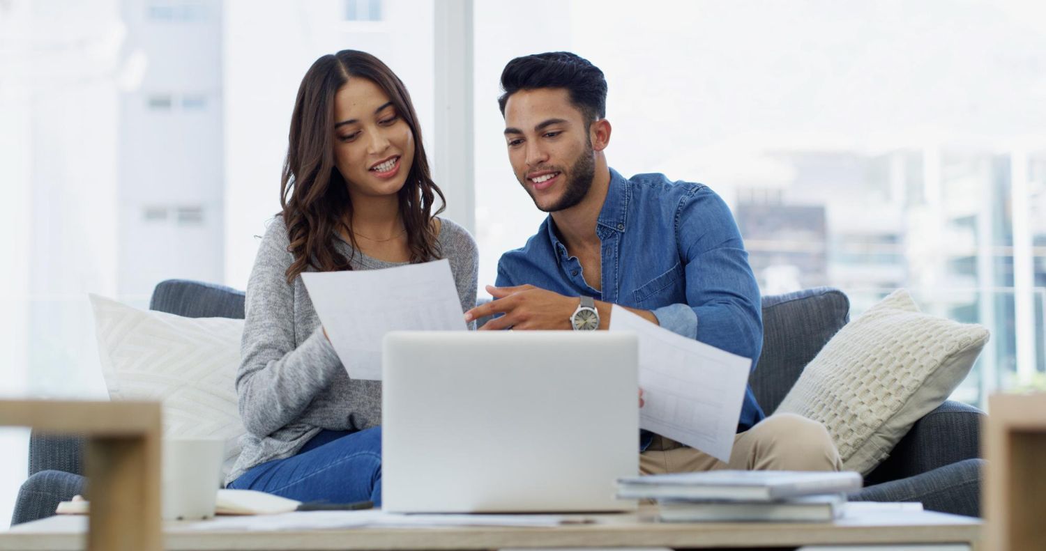 Couple reviewing paperwork, sitting on a sofa with a laptop. Smiling, in a brightly lit room.