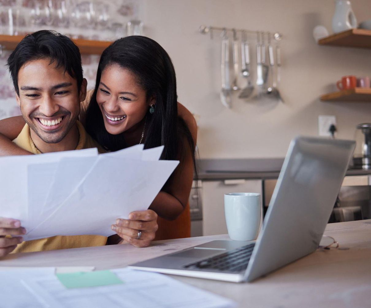 Couple smiling while reviewing papers together at a kitchen counter with a laptop.