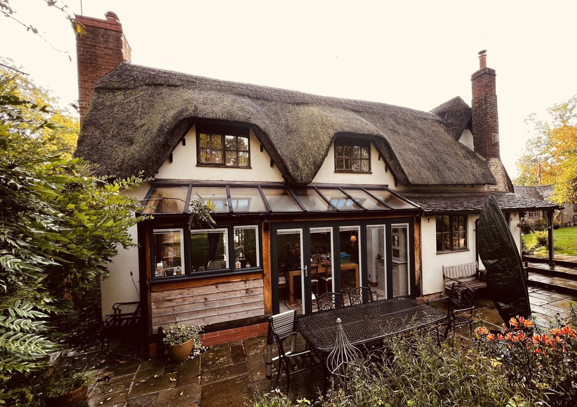 Cottage with a thatched roof, white walls, and a glass-walled extension. The setting is outdoors with trees and plants.