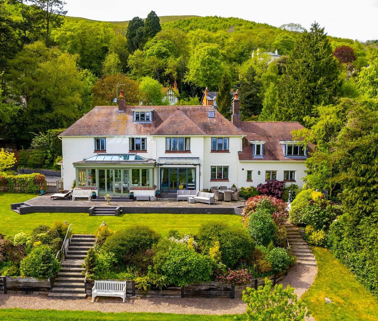 White house with a glass-walled extension, lush green garden, stone steps, and a bench. Surrounded by trees on a hillside.