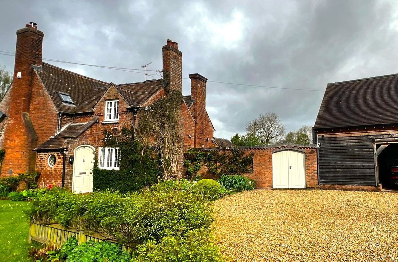 Brick cottage with chimneys, next to a gravel driveway and wooden barn, under a cloudy sky.
