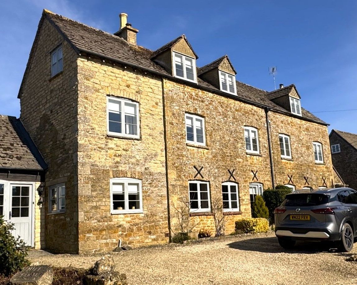 Stone house with white-framed windows, dormers, and a gravel driveway, with a car parked in front on a sunny day.