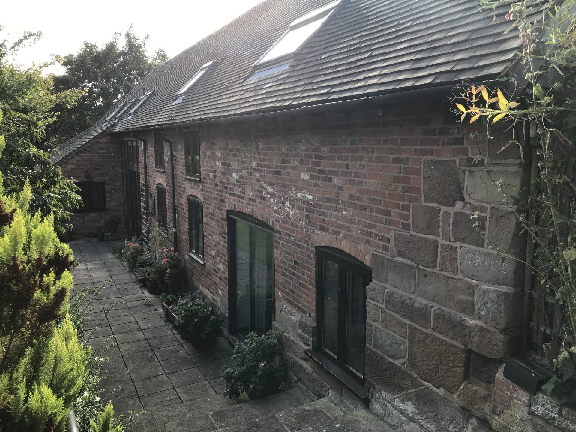 Brick building with dark roof and windows, fronted by stone walkway and greenery.