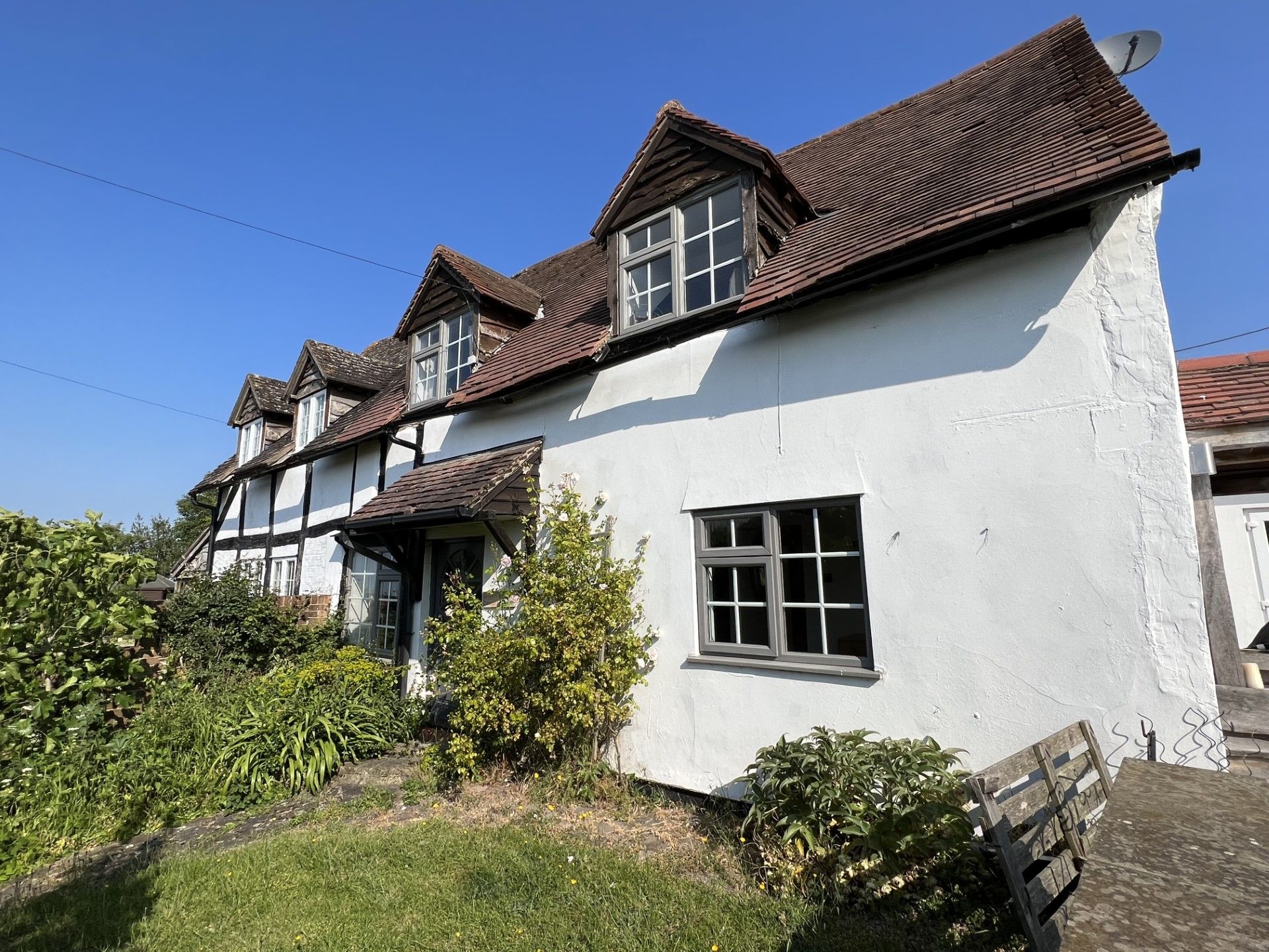 White cottage with timber beams, red tile roof, small windows, and overgrown garden under a blue sky.