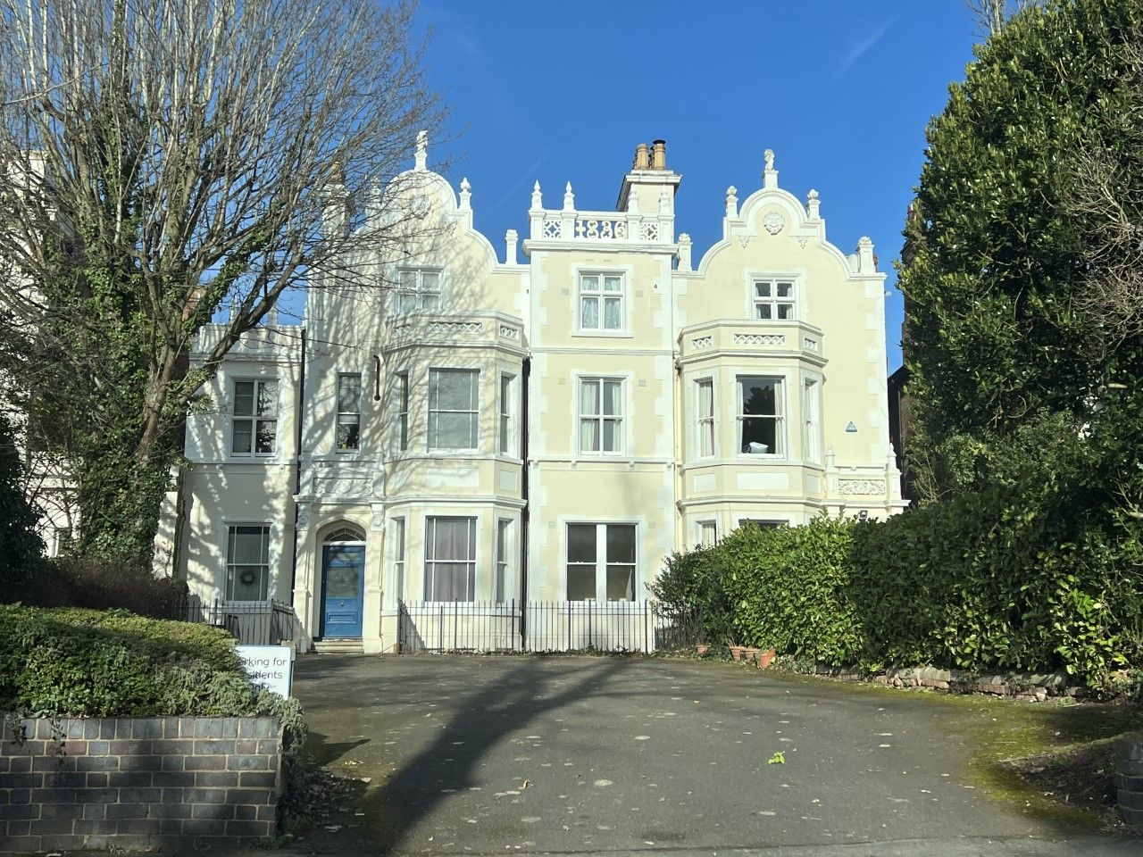 A light-colored building with ornate details, a blue door, and greenery on a sunny day.