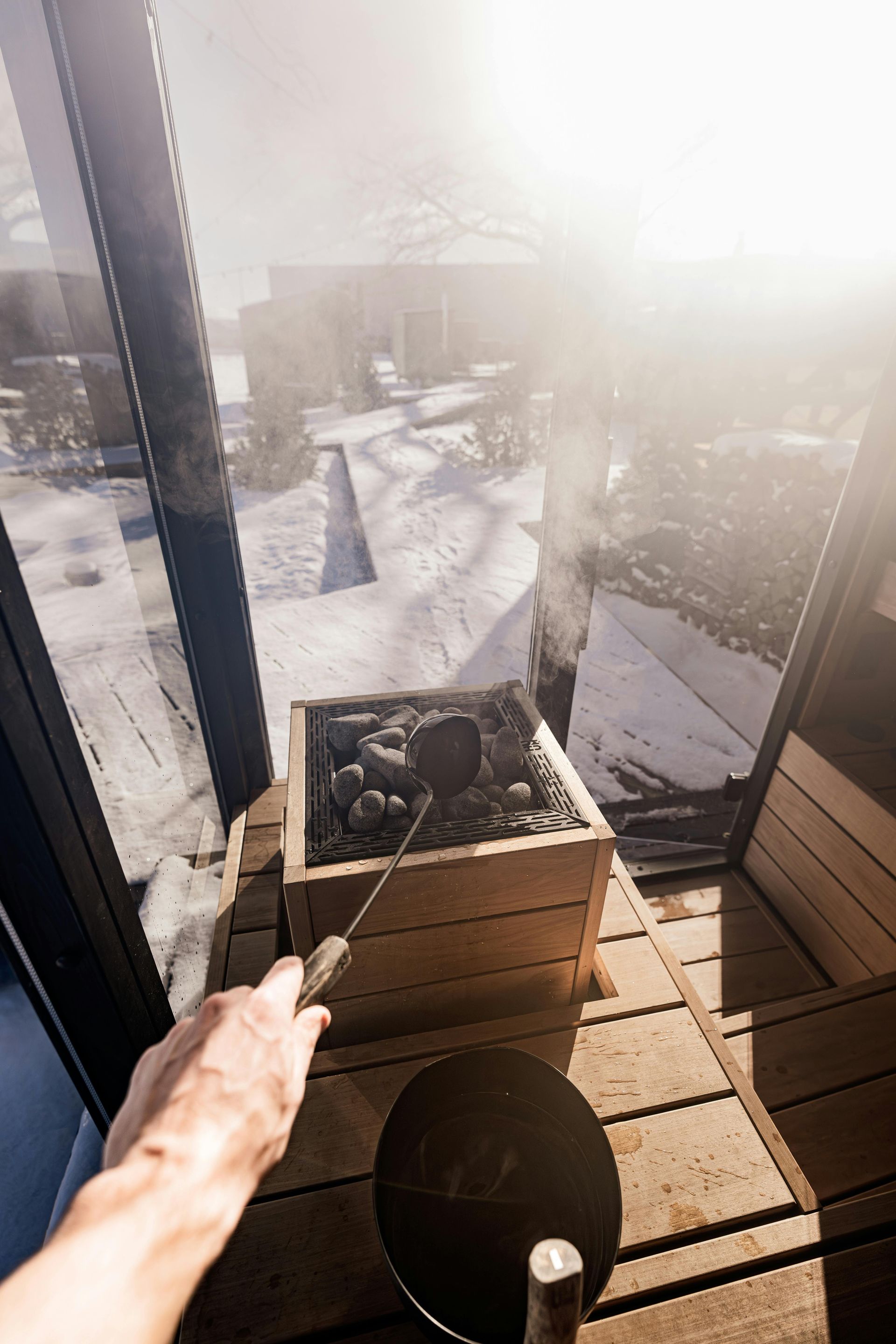 Balcone soleggiato con la neve fuori; una mano indica una pianta in vaso vicino alle porte a vetri.