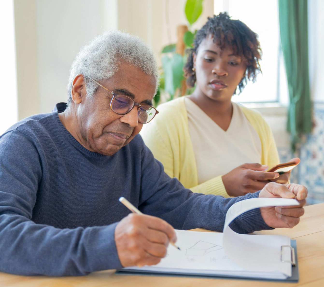 Older adult wearing glasses writing on a notepad, assisted by a person looking on and holding a phone.