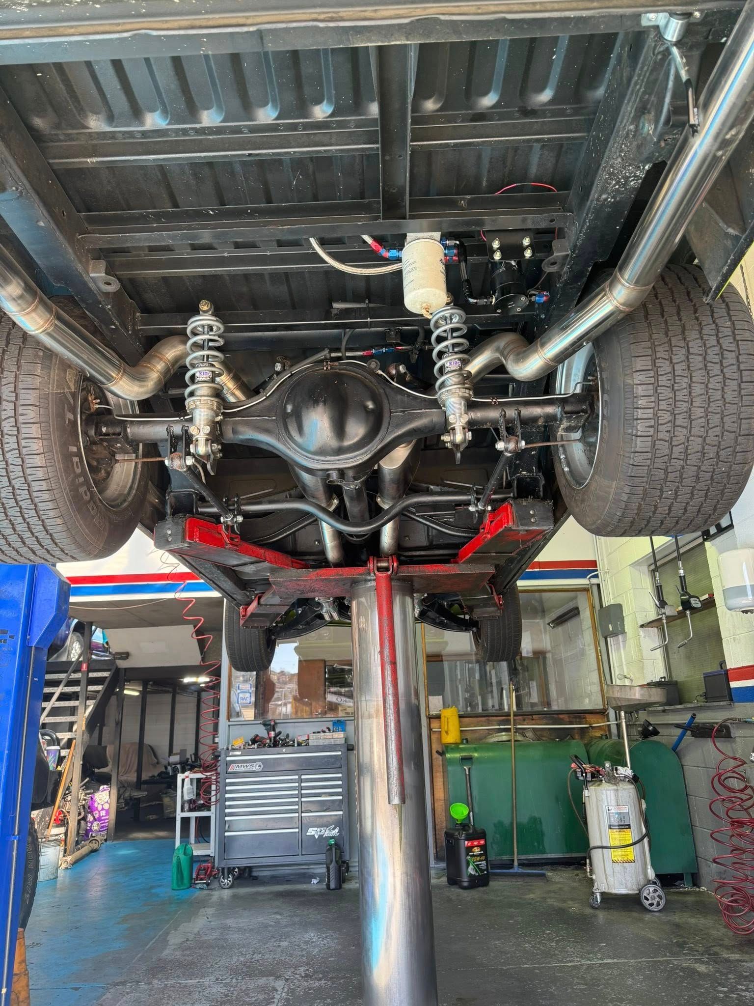 Undercarriage of a lifted vehicle on a shop hoist, showing suspension and exhaust in a garage