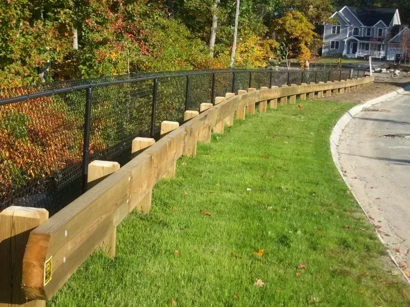 A wooden guardrail lines a curved roadside next to a black chain-link fence, with trees and a house in the background.