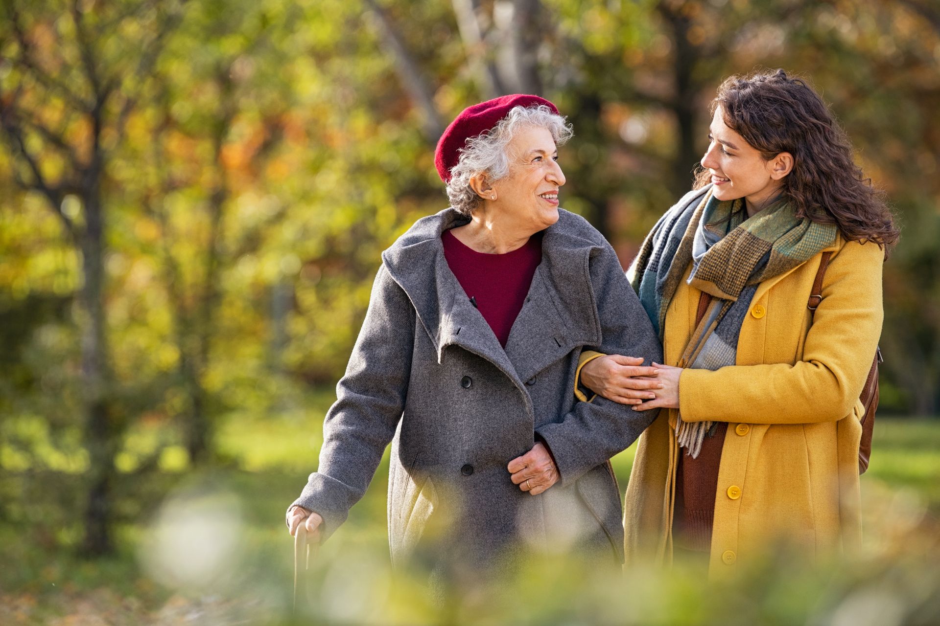 A person in a yellow coat walks with a person in a grey coat and a red beret in a park with autumn foliage.