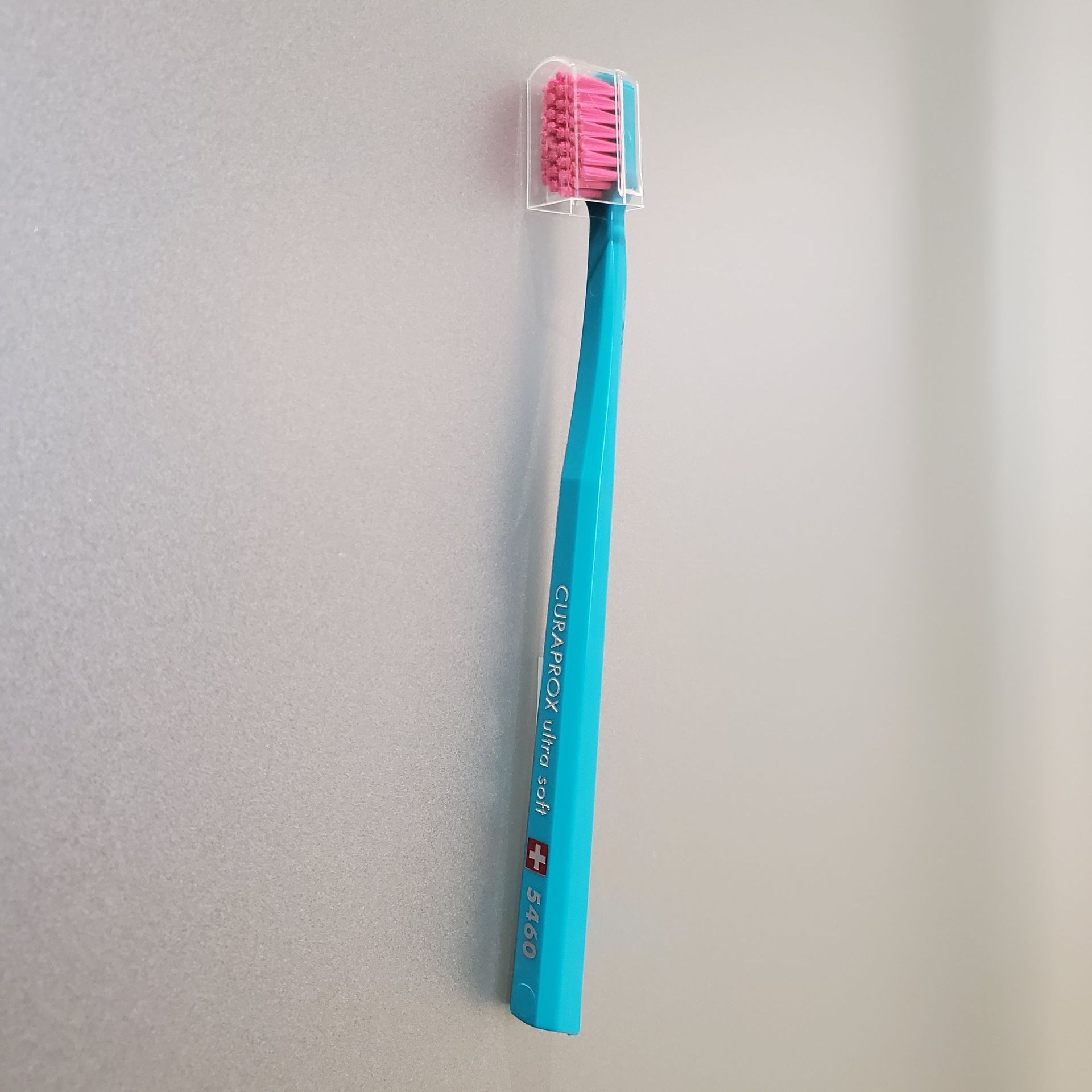 Blue toothbrush with pink bristles and a clear protective cover, against a light gray wall.