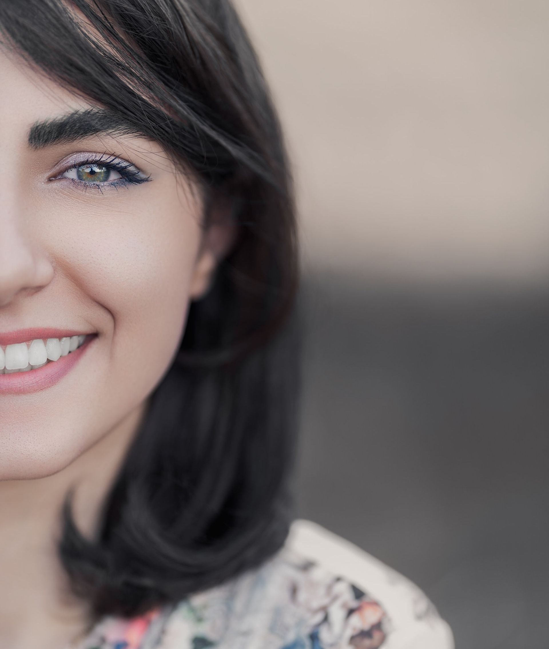 Woman smiles, half her face visible. Dark hair, sparkly eyeshadow, blurred neutral background.
