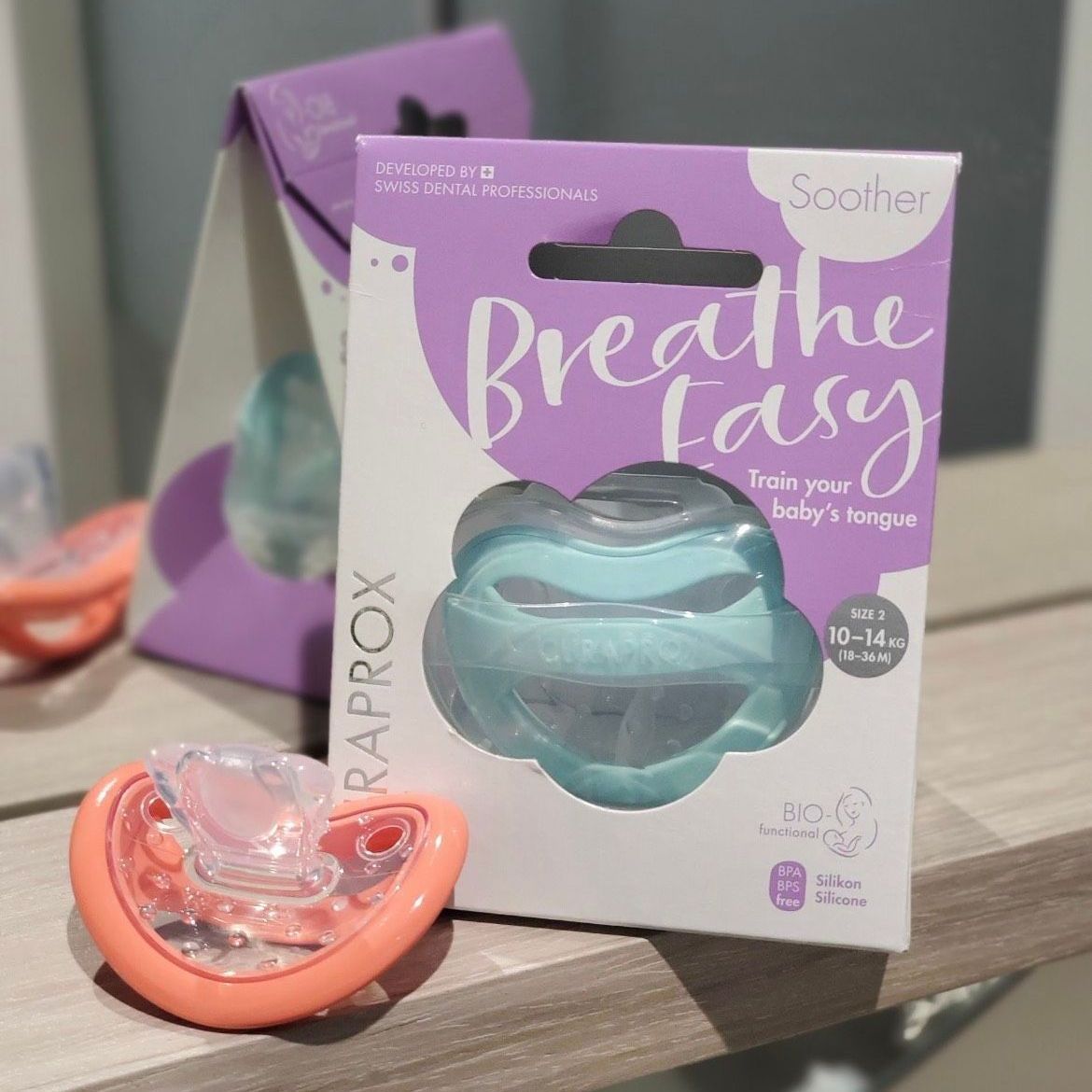 Pacifier box and pacifier. Box has “Breathe Easy” label. Pacifier is orange with a clear nipple.
