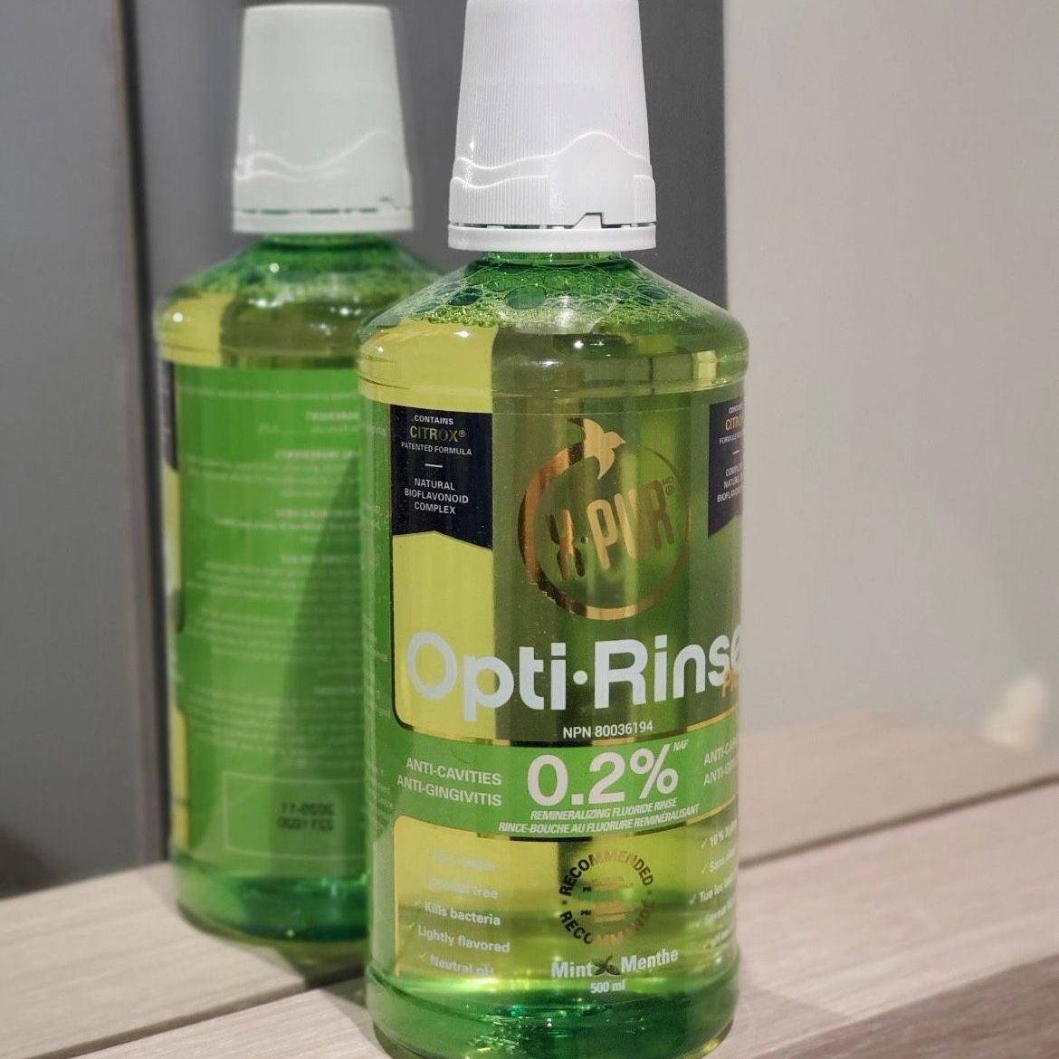 Two green bottles of Opti-Rinse mouthwash on a light wooden surface, with a green and yellow liquid.