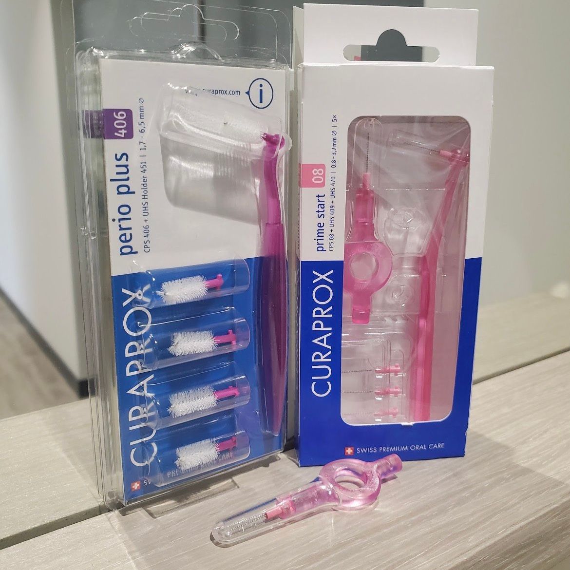 Two Curaprox interdental brushes, one package open, on a table. Pink and white bristles and handles.