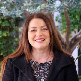 Woman with long brown hair smiles, wearing a black jacket, posing outdoors near a tree.