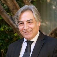 Man in a suit smiles, gray hair, outdoors near greenery.