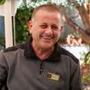 Man smiling, wearing a gray and black shirt. Outdoor setting with plants.