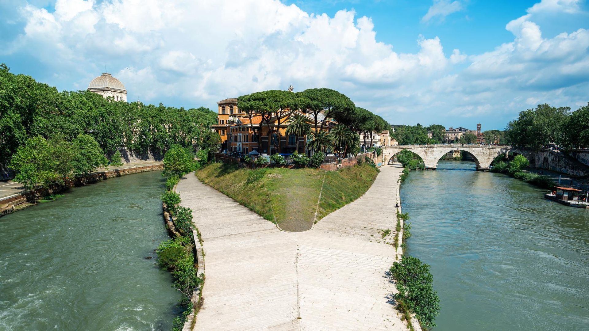 Isola del fiume Tevere con edifici e alberi, sentieri in pietra e ponti sotto un cielo azzurro.